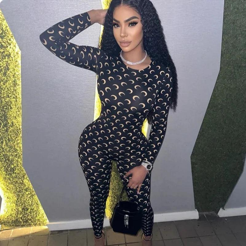 Woman wearing a black patterned outfit with a black handbag against a gray wall.
